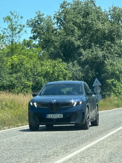 A dark blue BMW electric sedan is driving on a paved road in Debelets, Bulgaria. The car's front is visible, with its headlights on and a license plate reading "EA 8392 AE". The car is positioned on the right side of the road, with a white line marking the lane. The road is bordered by dry grass and a lush green forest in the background. The sky is clear and blue. A traffic sign with three circular discs is visible to the right of the car, partially obscured by trees. The overall scene depicts a daytime drive on a rural road in Bulgaria.
