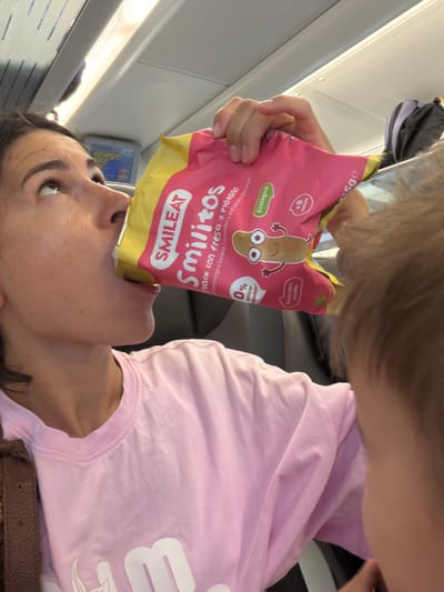 An adult woman wearing a pink t-shirt is shown from the chest up, leaning forward and drinking directly from a pink and yellow bag of SMILEAT "Smilítos" snack. The snack bag features an illustration of a cartoonish brown bean character with eyes and arms, and text in Spanish and English indicating it is a strawberry and banana corn snack with 0% added sugar and is ecological. The background appears to be the interior of a train, with a screen displaying a map visible above and to the left of the woman's head. A portion of a child's head and hair is visible in the foreground on the right side of the image, obscuring part of the woman's face and body. The overall lighting suggests an indoor, daytime environment. There is no indication of the city of Tarragona, Spain in this image.
