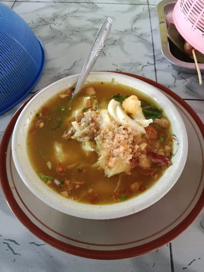 A close-up, high-angle shot captures a bowl of Suto (Soto) served on a light-colored, patterned tabletop in Sarimulyo, Indonesia. The central subject is a white bowl with a thin brown rim, placed on a larger beige plate with a brown inner rim. Inside the bowl is a light brown, opaque broth containing various ingredients: shredded white meat (likely chicken), pieces of hard-boiled egg with a visible orange yolk, finely chopped green herbs (possibly spring onion or celery), and small, reddish-brown crispy fried shallots or garlic flakes. A silver-colored metal spoon with an ornate handle rests vertically in the center of the bowl, partially submerged. To the left of the bowl, a portion of a blue, ridged plastic food cover is visible. To the right, a small rectangular metal tray holds a pink plastic basket with a domed lid, partially covering a green banana leaf and a single skewer holding a small, brown, elongated food item, possibly a sausage or fish cake. The tabletop features a subtle, wavy, light gray and white marbled pattern.