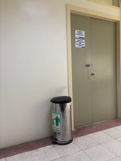 An indoor scene shows a light-colored wall on the left, a metal trash can on the right side of the wall, and a closed double door on the right. The trash can is cylindrical with a black lid and a foot pedal. A green label on the trash can features a pictogram of a person throwing trash and text in Arabic and English that reads "THE MEDICAL CITY" and "General Waste." The double door is a light olive green color and has a silver door handle and a bolt lock. Two white rectangular signs are affixed to the left side of the door. The top sign reads "NURSING SERVICE DEPARTMENT" and "Room No. 186." The bottom sign reads "HOSPITAL EPIDEMIOLOGY AND SURVEILLANCE UNIT." The floor is a terrazzo tile with speckled patterns and a narrow red border. The lighting suggests an indoor environment, possibly a hallway. There are no people present. The scene is forensic and matter-of-fact.