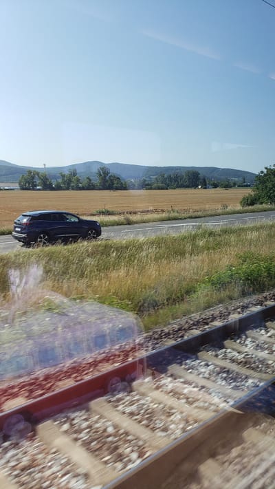 The image captures a roadside scene in Ivanovce, Slovakia, on a clear, sunny day. In the foreground, blurred train tracks run diagonally across the frame, with their gravel ballast and wooden sleepers visible. The reflection of what appears to be a train carriage is visible on the window, distorting the view of the tracks and the landscape beyond.

Further into the scene, a dark blue car, possibly a Peugeot 3008 based on its profile, is driving on a paved road that runs parallel to the train tracks. The car is viewed from its left side, and it appears to be moving. The road has clear white lane markings.

Beyond the road, a field of dried, golden-colored grass or crops stretches towards a line of trees and rolling hills in the background. The hills are covered in vegetation, and the sky is a clear, bright blue with a few faint wisps of clouds. A white building, possibly a house or small structure, is visible nestled amongst the trees on one of the hills. The overall impression is of a rural landscape on a bright summer day, viewed from a moving vehicle, likely a train, given the perspective and the presence of the train tracks.