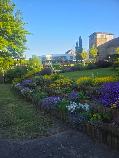 A vibrant outdoor scene in Görgeshausen, Germany, is depicted on a bright, sunny day. The image shows a meticulously maintained garden or park area, stretching horizontally across the midground. Prominent flower beds burst with color, featuring dense clusters of purple, yellow, and white blossoms arranged in distinct rows or sections. These flower beds border a neatly mown green lawn, which extends towards the background.

To the left of the lawn and flower beds, playground equipment with red and green frames is visible, suggesting a community play area. Lush green trees with full foliage dot the landscape, providing shade and natural beauty. In the background, under a clear, azure sky, stands a large stone building with a tower and a cross on its roof, indicating a church or chapel. Another building with a lighter facade can be seen further left, possibly a community center or residential structure. A dark, paved path or road runs along the right edge of the frame.

The weather is clear and sunny, with bright light illuminating the scene, creating vivid colors and clear visibility. A faint airplane contrail is visible in the distant sky. No people are present, and there is no visible text. The scene conveys a peaceful and well-kept environment, likely captured during mid-morning or early afternoon hours.