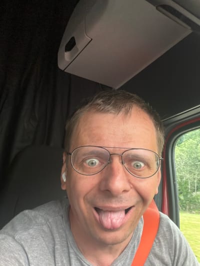 A light-skinned man with short brown hair and glasses is taking a selfie inside a vehicle. He is sticking his tongue out and has his eyes wide open. He is wearing a grey t-shirt and an orange seatbelt. The man is positioned in the foreground, with the interior of the vehicle visible behind him, including a black curtain and a grey sun visor. Outside the vehicle, through a window, a green, wooded landscape is visible. The specific location within the city of Peach Lake, United States of America, is not discernible from the image.