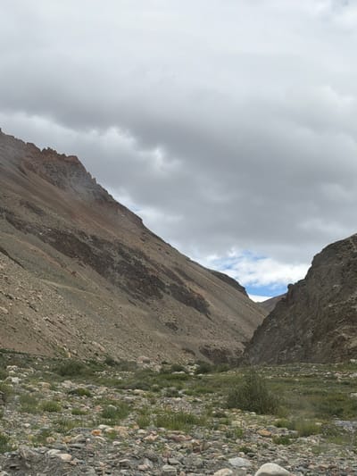 A wide-angle, eye-level, outdoor shot captures a rocky, arid valley under a cloudy sky. The rugged, sloped terrain dominates the frame, with the left side of the valley featuring a steep, multi-layered mountainside in shades of brown and gray. The right side of the valley presents a steeper, darker, and more shadowed cliff face. At the bottom of the valley, a rocky, gravelly ground is sparsely covered with low-lying green vegetation and small bushes. The scene appears to be located in the natural landscape of Kurgiakh, India, a region known for its mountainous terrain. There are no people or animals visible in the image.