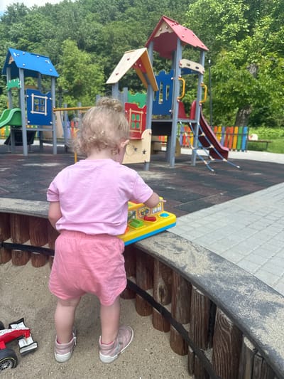 A blonde toddler with curly hair, seen from behind, is playing in a sandbox at a playground in Donchovci, Bulgaria. The child is wearing a pink t-shirt and pink shorts, and pink shoes. In front of the child is a yellow toy with colorful buttons and levers. A red and white toy car is visible to the left in the sandbox. The playground features a colorful slide, climbing structures, and a fence. The ground is a mix of rubber matting and paved tiles. The background consists of lush green trees.