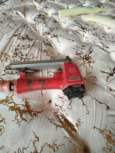 An upholstery project is underway, evidenced by a tufted fabric surface featuring a gold pattern. A red pneumatic staple gun lies on the fabric, suggesting ongoing work. Two pieces of foam padding are visible in the upper-right corner, ready for use. The scene indicates a process of furniture repair or creation, focusing on comfort and aesthetic appeal.
