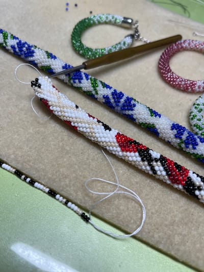 The image shows a close-up of several beaded items being created, likely jewelry such as bracelets or necklaces, and a crochet hook. The beaded items are tubular in shape and feature various colorful patterns. One has a pink and white pattern, another green and white, a third blue and white with green accents, and a fourth with a red, black, and white pattern. There are also loose beads scattered on the surface. The background is a textured, light-colored surface, possibly felt or a similar crafting mat, with a green surface visible on the right. A white thread is also visible, suggesting the beading process is in progress. The lighting is bright, indicating daytime, and there are no specific weather or location cues beyond the crafting materials. The focus is on the intricate beadwork and the tools used for its creation.