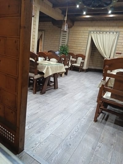 The image depicts an interior scene of a restaurant with a rustic aesthetic, likely in Derbent, Russia. The foreground is dominated by a wooden partition on the left and a wooden chair on the right. In the mid-ground, several wooden tables are set with light-colored tablecloths. The tables are surrounded by wooden chairs with upholstered seats and backs. The walls are constructed of light-colored brick, and a large, cream-colored curtain hangs on the right side of the frame, suggesting an alcove or entrance. The floor is made of light grey laminate wood planks with a prominent wood grain pattern. The ceiling features exposed wooden beams and recessed lighting, along with a decorative circular fixture that appears to be a fan. A hanging lamp with a wooden frame is suspended from the ceiling. The overall atmosphere is dimly lit and suggests an evening setting. There are no people visible in the image.