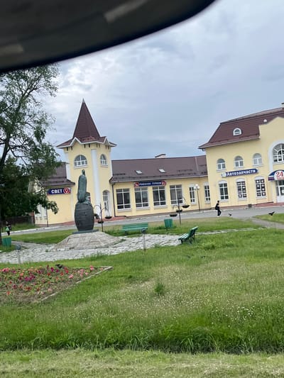 The image captures a scene in the city of Lukhovitsy, Russia, featuring a monument in the foreground and a yellow building complex in the background. The monument depicts a figure standing atop a barrel, sculpted in bronze. To the left of the monument, a sign reads "СВЕТ" (Svet), and to the right, a sign reads "РЫБАЛКА ТУРИЗМ" (Fishing Tourism). The building complex appears to house several establishments, including one with a sign reading "Автозапчасти" (Auto Parts) and another with "24 часа" (24 Hours) indicating round-the-clock service. Green park benches are visible in the grassy area between the monument and the buildings. A lone person in dark clothing walks across the paved area in front of the establishments, with two birds on the ground nearby. The sky is overcast with clouds, suggesting a temperate climate. The overall setting appears to be a public square or a street view of commercial buildings with a cultural monument.
