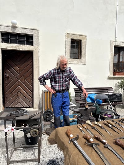 An outdoor scene in Častá, Slovakia, captures an elderly man, likely a blacksmith, at what appears to be a market stall or open-air workshop. The man, with long white hair and wearing a red and black plaid shirt over blue trousers, stands next to a large anvil. He holds a hammer, poised as if working or demonstrating.
To his right, a brown cloth draped over a surface displays an array of handcrafted knives and swords with various handle designs made from wood and bone. Further right and below the anvil, a forge or furnace setup with an electric blower is visible, along with blacksmithing tools and coal. A small white and red ribbon is tied to the forge's stand.
The background features a white stucco building with three windows, two with dark frames and bars, and a large, intricately designed wooden door with a geometric pattern. A wooden bench is situated to the right of the man. The bright, clear lighting suggests a sunny day, with soft shadows indicating morning or afternoon. The overall setting evokes a traditional craft demonstration or a local market showcasing artisan goods. A numerical label "30" is visible on the side of the anvil.