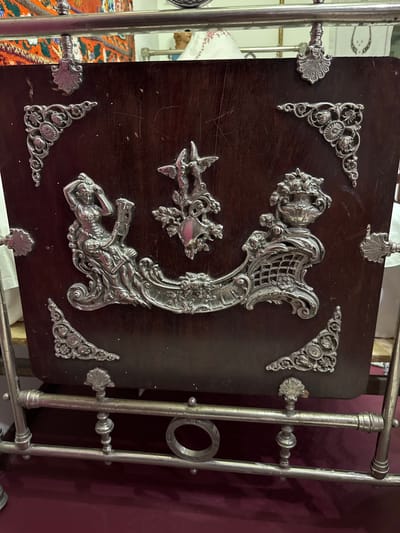 The image shows a close-up view of an ornate, decorative object, possibly a fireplace screen or a decorative panel. It is made of dark wood with intricate silver-colored metal embellishments. These embellishments include floral motifs, corner decorations, and a central scene depicting a figure of a woman holding a scroll or book, a winged cherub, and a decorative vase with floral arrangements. The object is supported by a metal frame, which appears to be made of silver or a similar polished metal. The background is blurred but suggests an indoor setting with textiles and other objects visible, hinting at an antique shop or a museum display. The object is located in Fort-Şevtşenko, Kazakhstan.