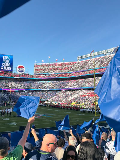 A vibrant stadium filled with fans dominates the frame, the stands a mix of red, white, and blue. A "Pepsi" sign and a screen reading "RAISE YOUR CARDS & FLAGS" are visible. In the foreground, spectators wave blue flags, obscuring parts of the scene. The clear blue sky above contrasts with the colorful energy of the stadium crowd.