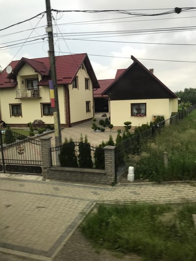 This image depicts a residential scene in Vicovu de Sus, Romania, under an overcast sky. The foreground features a light-colored interlocking paver pathway or section of road, bordered by a darker stripe, which transitions into an unkempt grassy area on the right.

The midground is dominated by a private property entrance. A dark metal gate with decorative scrollwork is positioned on the left, flanked by two stone pillars constructed from light-colored bricks. A matching metal fence extends to the right, supported by similar stone pillars and a low brick wall. Beyond the gate, a paved driveway, mirroring the foreground's interlocking pavers, curves between two houses. The property's landscaping includes several manicured evergreen shrubs, some shaped into conical or spherical forms, arranged in garden beds and along the driveway.

A tall utility pole stands prominently in the center of the image, extending upwards with numerous black overhead wires crisscrossing against the grey sky. A rectangular grey utility box is attached to the pole, from which a vertical banner displaying the blue, yellow, and red stripes of the Romanian flag hangs.

The primary structures are two houses, both featuring light yellow stucco exteriors and red tiled roofs. The house on the left is a two-story building with multiple gables. A silver satellite dish is mounted on its left roof section. The facade includes dark-framed windows, some with window boxes containing red flowers. A balcony with a silver metal railing projects from the upper floor. Decorative dark brown trim outlines the windows and corners, the latter also featuring vertical decorative brick-like elements. A person, partially visible and seemingly crouched or kneeling, is situated near the lower left side of this house.

The house on the right, partially visible, also has light yellow walls and a red tile roof. Its most striking feature is a prominent dark brown, wood-paneled gable end facing the viewer. A dark-framed window with a window box of flowers is visible on this facade. To its left, a recessed garage or covered area is visible, containing a small red and black ride-on toy.

In the background, an elevated grassy slope with scattered vegetation runs along the right side of the frame, bordered by a simple fence. The sky is uniformly overcast.