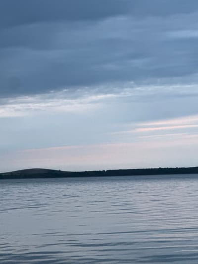 The image shows a body of water with ripples, likely a lake or river. The water is a muted blue-grey color, reflecting the sky. On the left side of the frame, the sky is a soft blue with streaks of white clouds. Towards the right, the sky transitions to a paler shade, with hints of pink and orange, suggesting either sunrise or sunset. A dark, silhouetted treeline or landmass forms a thin line separating the water from the sky on the right side of the image. The overall mood is calm and serene, with the soft lighting and natural elements. The time of day is likely early morning or late evening, given the sky colors. The location cues, if interpreted as land, are minimal, but the presence of a large body of water and a distant tree line are visible. No people or objects are discernible in the image, and there is no visible text.