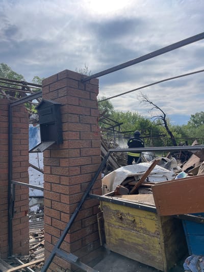 The image shows a partially destroyed brick building with a black mailbox attached to the side. The roof has been completely destroyed, with a large amount of debris scattered throughout the structure. A person in a black uniform with a fire extinguisher is visible in the background, working among the debris. The location is a residential area in the city of Bilopillia, Ukraine, with coordinates 51.139 latitude and 34.285 longitude. The sky is cloudy with a few bright spots, and the environment is dry and dusty. The scene suggests a recent fire and the aftermath of the event.