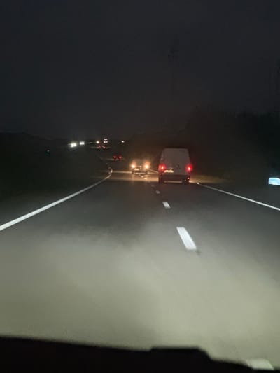 Nighttime scene on a highway. A large truck with illuminated red tail lights is visible ahead in the lane. Several other vehicles, indicated by their headlights and taillights, are also in the distance. The road is marked with white lane dividers. The right side of the frame shows a brighter concrete surface. Dark, undefined areas frame the left and right edges of the image.