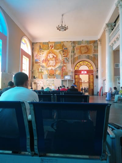 The image depicts the interior of a waiting area, likely a train station or airport terminal, in Dubrovka, Russia. The scene is filled with people sitting on dark blue chairs, facing away from the camera. The most prominent subject is a man in the foreground, wearing a white t-shirt, seated with his back to the viewer.
The waiting area features a large, colorful mural on the back wall, depicting historical or mythological figures and maps. Above the mural, a decorative chandelier hangs from the ceiling. A large arched doorway with an illuminated clock showing "30.06" is visible in the center of the back wall. The architecture includes tall, fluted columns and arched windows.
Several people are scattered throughout the waiting area, some engaged in conversations, others looking at their phones or resting. The lighting suggests it is daytime, with natural light filtering in from the windows. The overall atmosphere appears to be one of quiet anticipation, typical of a travel hub.
The visible text includes "30.06" on the clock. There is also some illegible text on what appears to be a bulletin board on the back wall, and some scribbled numbers on the floor in front of the chairs. The presence of a large mural with historical imagery and Cyrillic text suggests a location in Russia. The specific mention of "Dubrovka" further grounds the location.