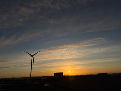 This image depicts an outdoor scene at sunrise or sunset over Naaldwijk, Netherlands. The lower third of the frame presents a dark, silhouetted landscape composed of numerous low-rise urban and industrial buildings, indistinct structures, and sparse vegetation. Patches of what appear to be wet ground are visible in the immediate foreground. A large, three-bladed wind turbine, with blades positioned as if stationary or slowly turning, stands prominently silhouetted against the sky on the left side. The sun, a bright circular orange-yellow orb, is positioned low in the sky, slightly right of the image center. The sky occupies the upper two-thirds of the image, displaying a gradient from deep blue-grey at the top, transitioning through lighter blues, to vibrant orange and yellow hues near the horizon. Multiple layers of scattered cirrus and altocumulus cloud formations are present, along with at least three distinct, linear white aircraft contrails visible across the sky. No human figures or specific actions are discernible within the frame.