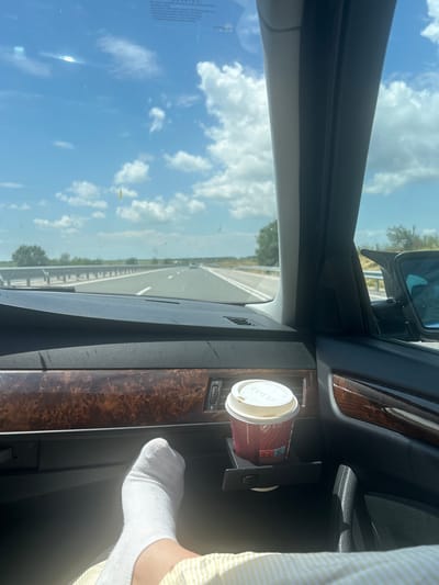The image depicts the interior of a car from the passenger's perspective, traveling on a multi-lane highway in Yazdach, Bulgaria. The sky overhead is bright blue with scattered white cumulus clouds, indicating fair weather. The car's windshield is visible, showing some dirt, streaks, and a small yellow object adhered to it, possibly a leaf or insect. A light-colored vehicle is seen in the distance on the highway, moving in the same direction. Green trees and foliage line the sides of the highway, receding into the background.
Inside the vehicle, a dark-colored dashboard with a textured surface is prominent, accented by a polished, reddish-brown wood trim panel featuring a swirled grain pattern. A person's left foot, clad in a white sock and part of a yellow and white striped garment, rests casually on the dashboard area to the right of the glove compartment. Adjacent to the foot, a red Costa Coffee disposable cup with a white lid is secured in a pull-out cup holder integrated into the dashboard. The car's dark interior A-pillar and a portion of the passenger door with its handle are visible, along with the right side mirror reflecting part of the external environment. A faint "WARNING!" label or similar text is reflected on the upper part of the windshield.