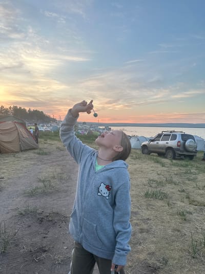 A young person with blonde hair pulled back is standing in a field with dry grass and dirt. They are wearing a gray hooded sweatshirt with a Hello Kitty design and dark pants. The person is looking up and holding a blue, round object on a string to their mouth. In the background, there is a body of water, a shoreline with trees, and a campsite with several tents and a silver SUV. The sky is a soft blend of blue, pink, and orange, suggesting it is either sunrise or sunset. This scene is likely in the city of Zhemchuzhny, Russia, given the presence of the campsite and natural landscape.