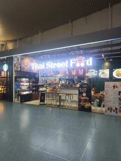 The scene depicts an interior view of a commercial food court or terminal in Tambon Sa Khu, Thailand, characterized by high ceilings and a grey speckled floor. In the foreground, a prominent glass-fronted "Thai Street Food" restaurant occupies the center-right, featuring a brightly lit sign with the restaurant's name and a red food cart logo. To the left of this restaurant, a Starbucks coffee shop is partially visible, identifiable by its green circular logo and a display case with baked goods.
Inside the "Thai Street Food" establishment, several elements are discernible:
* **Frontage:** The entrance is marked by a yellow and black striped tape on the floor.
* **Displays:** A refrigerated display unit containing bottled beverages and other items is positioned near the Starbucks boundary. Further inside, multiple display cases showcase pastries and various food items.
* **Counter and Seating:** A light wooden counter serves as the service area, behind which at least two staff members are present. Seating arrangements include small wooden tables with white chairs. One customer is seated at a table toward the back right.
* **Signage:** Large digital menu boards display images of Thai dishes, burgers, and bubble tea. A standalone black A-frame sign promotes "O2S Burgers & Beer" with an image of a burger and fries. An additional vertical menu board with text and drink images is on the far right.
* **Decor:** The interior features a dark green wall paneling on the right side with decorative sconces.
The environment is brightly lit by overhead strip lighting, with reflections visible on the polished floor. The overall impression is that of a clean, well-maintained, and active commercial food service area within a modern facility.