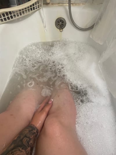 The image shows a person's legs submerged in a bathtub filled with foamy water. The scene is set in a bathroom, indicated by the presence of a bathtub, a drain, and a shower head with a flexible hose. A plastic laundry basket is partially visible on the left side of the tub, suggesting domestic use. The water is cloudy, with a significant amount of bubbles covering the surface. The person's hand is visible in the water, with a manicured fingernail and a tattoo on their forearm. The lighting suggests an indoor setting, possibly during the day, but no specific time of day or weather conditions are evident. The overall mood appears relaxed, as one might expect during a bath. There is no visible text in the image.