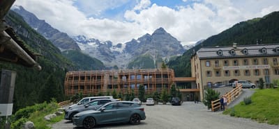 This is a photograph of the Bella Vista Hotel, also known as "Schöne Aussicht," located in Stilfs - Stelvio, Italy. The hotel is a multi-story building with a light-colored facade and a dark-tiled roof. It features numerous windows with shutters and balconies. In front of the hotel, a modern, multi-story building with a wooden facade and balconies is visible.

The hotel is situated in a mountainous region, with lush green slopes covered in coniferous trees rising behind it. Majestic, snow-capped mountains dominate the background, with a prominent peak in the center. The sky is partly cloudy with patches of blue.

In the foreground, a parking lot is visible with several cars parked, including a dark grey hatchback, a white van, and a silver SUV. Two individuals, a male and a female, are standing on a wooden railing that separates the parking area from a grassy slope. The male is wearing a pink shirt and dark shorts, while the female is wearing a light blue shirt and dark shorts.

The scene depicts a scenic mountain resort with modern architecture blending with the natural landscape.