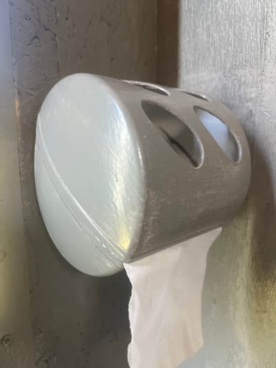 In Tashelka, Russia, a light gray toilet paper dispenser, mounted on a textured gray wall, dispenses a sheet of white toilet paper. The dispenser is made of metal and has an oval shape with three large circular cutouts along its upper surface. The wall appears to be constructed of plywood or particle board, with a visible grain pattern. The lighting is dim, creating shadows and highlighting the reflective surface of the dispenser. No people are present in the image.