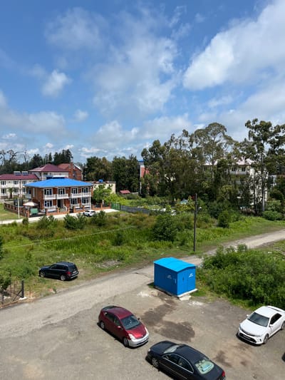 This image depicts an outdoor scene in Ureki, Georgia. The setting appears to be a street or road adjacent to a residential area with multiple buildings and lush greenery. The sky is visible with scattered white clouds against a blue backdrop, suggesting daytime with fair weather.
Several objects are present, including a bright blue electrical transformer box situated on the side of the road. Various cars are parked or driving on the asphalt road: a white car, a black car, and a red and silver car are clearly visible. The buildings in the background vary in architectural style, with some featuring red tiled roofs and others with blue roofs. Some buildings have multiple balconies. Tall trees and dense vegetation line the road, contributing to a natural, somewhat rural or suburban feel.
There are no people clearly visible in the image. The image is taken from an elevated perspective, looking down at an angle. The overall impression is one of a calm, everyday scene in a town or village. No discernible text is visible in the image.