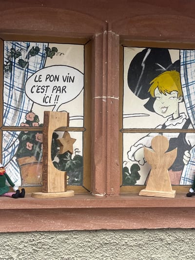 The scene depicts a two-pane window display on a building in Kintzheim, France. The window features reddish-brown stone framing and a central mullion, with a matching stone windowsill below. The wall beneath the sill is light-colored and textured.
Behind the glass, each window pane displays a painted backdrop. The left pane shows a blue and white checkered curtain, a potted plant with pink flowers, and green foliage. A white speech bubble is painted in the upper section, containing the French text "LE BON VIN C'EST PAR ICI !!" (The good wine is here!!). The right pane depicts a cartoon character with bright yellow hair, a smirking expression, a ruffled white collar, and a black hat or garment. A blue and white checkered curtain is partially visible on the far right of this pane, and green foliage is also present. The character's right hand is visible and appears to be pointing towards the left, consistent with the speech bubble's direction.
On the reddish-brown windowsill, several objects are arranged. On the far left, a small elf doll, clad in green and red-and-white striped attire with black shoes, sits facing slightly right. Centered between the panes is a natural wood sculpture with a raw bark edge on one side and a smooth, curved cut-out in its body; a five-pointed wooden star is suspended within this cut-out. To the right of the central mullion, a light-colored wooden silhouette of an angel stands upright on a flat base. A second elf doll, similar to the first, is partially visible on the far right of the windowsill, with only its green leg and black shoe discernible.