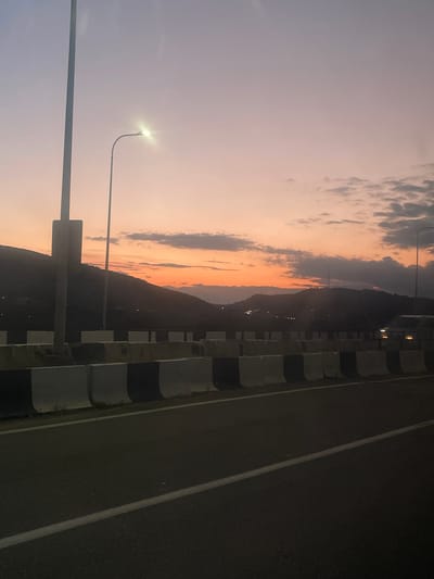 An outdoor, wide-angle, eye-level shot shows a road in Changilari, Georgia during a sunset. The sky is a gradient of purple, pink, and orange, with scattered clouds. In the foreground, a paved road runs diagonally across the frame, marked with a white line. To the left, a concrete barrier with alternating black and white sections separates the road from a steep, dark hillside. A tall, grey lamppost stands near the left edge of the frame, and another lamppost with a lit bulb is visible in the mid-ground against the colorful sky. Distant mountains are silhouetted against the horizon, with a few lights visible on their slopes, suggesting a settlement. A vehicle's headlights are visible in the distance on the right side of the frame. The overall atmosphere is serene and tranquil.