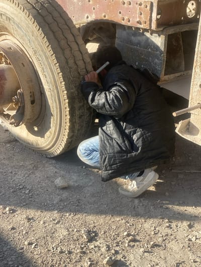 In Dahiyat Harasta, Syria, a single individual is observed in a crouched position outdoors, inspecting the underside of a large, stationary vehicle. The person, viewed from the back, wears a black quilted jacket, blue denim jeans, and white athletic shoes. They are holding a silver, cylindrical object, consistent with a flashlight or inspection tool, and directing it into the space between a large, dirty tire and the vehicle's heavily corroded metal chassis. The vehicle's frame exhibits numerous pockmarks, consistent with impacts or severe material degradation, and rust is prevalent. Exposed rebar or similar metal rods are visible extending from a structural component on the far right. The ground beneath is composed of loose dirt and small gravel, illuminated by strong daylight casting distinct shadows.