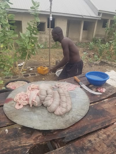 The image depicts a man standing in front of a partially constructed building in Pati Ridge, Nigeria. He is wearing black shorts and is shirtless. His back is to the camera and he appears to be reaching down towards the ground. A large piece of raw meat is on a metal plate resting on top of a wooden table. A knife is lying beside the plate, along with a blue bowl and other pieces of meat. The surrounding area is dusty and overgrown with small plants. The scene likely takes place outdoors.  There is a partially constructed building in the background with a lightbulb hanging on a wire.  There are other pieces of meat in a black container in the background as well.