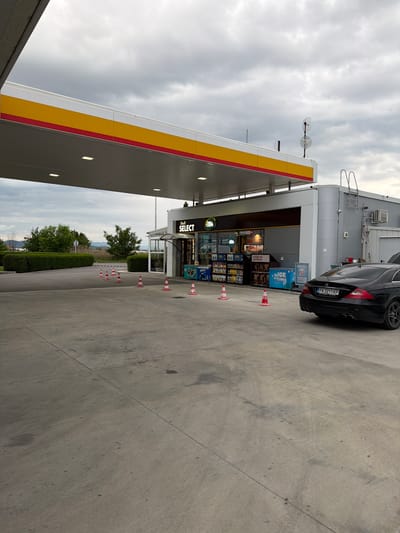 The image depicts a Shell gas station in Dragor, Bulgaria. A black Mercedes-Benz is parked next to the convenience store. There are several traffic cones lining the pathway between the store and the road. The sky is overcast.