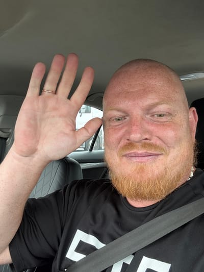 In a close-up selfie taken from inside a vehicle, a bald, fair-skinned man with a ginger beard and mustache is visible. He is wearing a black t-shirt with a white logo and a seatbelt. The man is smiling and waving with his left hand, which has a ring on the ring finger. The background shows the interior of the car, including a grey headliner and a black quilted seat. Outside the car window, a white truck is partially visible. The image appears to have been taken in Zanevka, Russia.