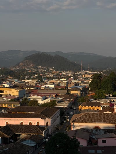 This image shows an elevated, long-range view of the city of La Esperanza, Honduras. In the foreground is a close collection of buildings with roofs of varying styles and colors. Many of the structures are pastel shades of pink, yellow, and white. Further into the distance, there is dense urban development. The roofs of the buildings are densely packed and varied in color. There is a large mountainous landscape to the side of the city. The sky is hazy, and there appear to be few clouds. A communications tower stands out as a tall, narrow structure among the other buildings. There is an active street in between buildings and cars driving on it.