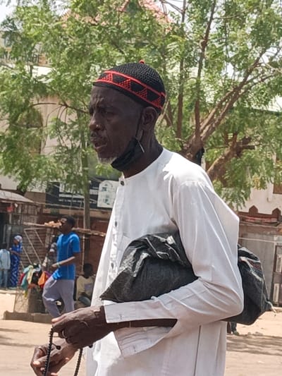 The image depicts an elderly Black male subject in the city of Hotoro, Nigeria, positioned in the foreground and facing left. He wears a black knitted skullcap with a red geometric pattern, a white long-sleeved tunic, and a black face mask suspended below his chin. His hands hold a string of black prayer beads, and he carries a dark grey or black plastic bag tucked under his left arm. His mouth is slightly open. The background reveals an outdoor, urban environment under bright daylight. Green-leafed trees dominate the upper background. In the midground, several indistinct figures are visible, including a man in a blue t-shirt and light-colored pants walking left, and a person in a blue patterned dress. Informal structures, possibly market stalls or residential buildings, are present in the midground and further back. The ground appears unpaved.