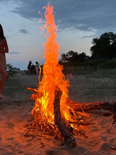 A nighttime scene shows a large bonfire on a sandy beach, likely in Novo Oriahovo, Bulgaria. The fire is built around a large piece of driftwood. Flames rise high into the air against a backdrop of a twilit sky and distant foliage. Three people are silhouetted against the sky and are standing near a grassy dune in the distance. A person is seated in the foreground, at the bottom of the image, with a visible upper arm and a cup in hand. The foreground is the sandy beach.