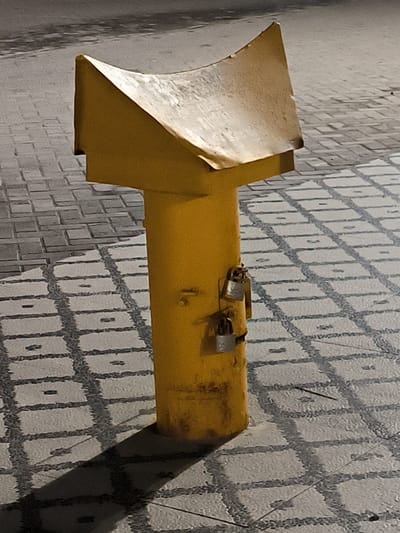 A yellow, metal, T-shaped object stands on a gray, patterned pavement. The object has two padlocks attached to its cylindrical portion. The pavement is composed of square tiles with darker grout. The location is Kotapantonlabu, Indonesia. The light source is above and to the right, casting a shadow from the object on the pavement.