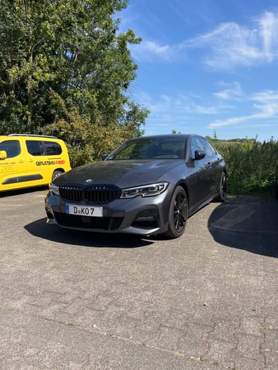 A dark gray BMW station wagon is parked on a paved area, adjacent to a bright yellow van. The BMW is positioned slightly in front of and to the right of the van. The car has a black grille and a license plate that reads "D-KO7". The van has text on its side that appears to be "GERUSTBAU KAISER" along with an address and phone number. The scene is outdoors, with greenery and trees visible behind the vehicles. The sky is blue with scattered white clouds. The lighting suggests it is daytime, likely sunny given the shadows cast by the car and the bright colors. The ground is a light gray paved surface made of interlocking stones. The overall impression is a utilitarian parking or work area. There are no people visible in the image.