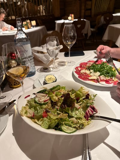 A restaurant table setting features a vibrant salad, a plate of carpaccio with greens and Parmesan, and a small dish of cheese. Water and wine glasses flank the dishes, with Christmas ornaments visible through the wine glass. Cutlery is placed near the salads and carpaccio. The scene is illuminated by soft, ambient light, highlighting the colors and textures of the food.