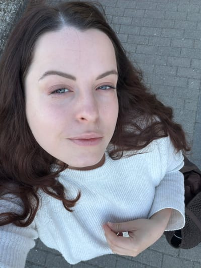 A woman with light skin, dark brown hair, and blue eyes is pictured from a low-angle perspective. She is wearing a white, textured knit sweater. Her hair is spread around her head, and her right hand is resting near her right shoulder. The background shows a paved surface, likely a walkway or street, with dark brown fabric. There is insufficient information to conclude her location in Odernheim am Glan.