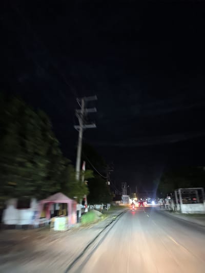 This is a night-time, first-person perspective photograph taken from a moving vehicle on a paved road in Keudeteunom, Indonesia. The image captures a street scene with several elements. On the left side of the road, there is a small, pink stall or kiosk with some items displayed, and behind it, a building with a white facade. Adjacent to the stall are trees, and a utility pole with electrical wires stands tall. Further down the road, there are more trees and buildings visible. The road itself is illuminated by the headlights of the moving vehicle and the lights of other vehicles ahead. Two motorcycles with red taillights and a car with illuminated headlights are on the road. The sky is dark, with some faint cloud formations. The overall impression is one of a rural or semi-urban street at night, with typical infrastructure and light traffic.