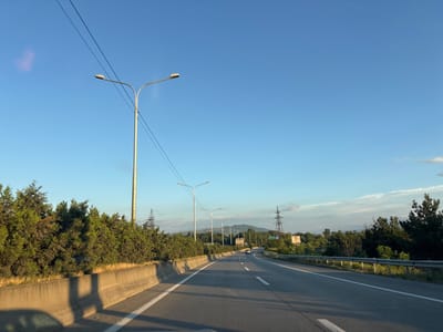 A wide, grey asphalt highway curves gently to the right under a clear, pale blue sky. The highway has two lanes in each direction, separated by a dashed white line. Streetlights on tall poles line the right side of the highway, casting long shadows onto the road. A concrete barrier runs along the left side of the road, bordered by lush green trees and bushes. In the distance, rolling hills and mountains are visible, some topped with power line pylons. The sun appears to be setting, as evidenced by the long shadows and warm light illuminating the scene. The overall atmosphere is peaceful and serene, with a sense of open road and travel. There is no visible text in the image.
