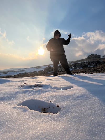 This image depicts an outdoor, snow-covered landscape in Talfita, Syria, under a bright, partly cloudy sky. A single individual, dressed in a dark hooded sweatshirt and dark pants, stands in the right-center of the frame, largely silhouetted against the low sun on the left horizon. The person's posture, with legs apart and arms raised, suggests an active motion; a small, spherical white object, consistent with a snowball, is visible in the left hand, while the right hand is open with visible water droplets around it, indicative of melting snow or ice. The immediate foreground features pristine snow with a prominent depression revealing dark soil and sparse, dry plant stalks. The midground consists of a gentle incline covered in snow and scattered piles of rocks. In the distant background, atop a hill to the right, a multi-story, seemingly abandoned or unfinished building with dark, empty window openings is visible. Long shadows are cast across the snow from the figure and other elements, illuminated by the strong backlighting from the sun, which also produces a visible lens flare.