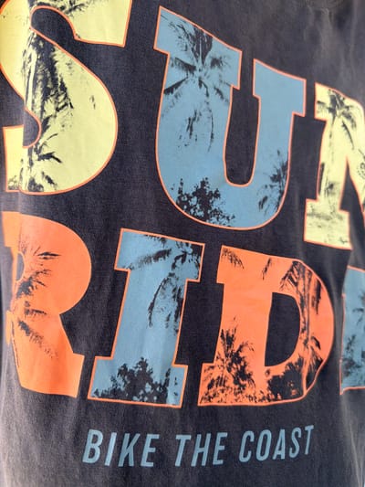 A close-up, low-angle shot shows a dark gray t-shirt with large, stylized text and graphics. The text "SUN RIDE" is printed across the chest of the t-shirt in various colors, including yellow, light blue, and orange, each with an orange outline. Within the letters, the graphic appears to be a silhouetted palm tree fronds. Below "SUN RIDE," the text "BIKE THE COAST" is printed in a light blue color. The t-shirt is made of a textured fabric, likely cotton. The image does not contain any information about a location in Dinkata, Bulgaria.