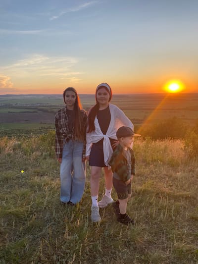 Three children are standing on a grassy hill overlooking a landscape of fields and a small village at sunset. The child on the left is a girl with long, dark hair, wearing a plaid shirt tied at the waist and light blue jeans. The child in the middle is a girl with dark hair and a headband, wearing a white cardigan over a black top and a dark skirt, with white sneakers. The child on the right is a young boy wearing a cap, a green and black plaid jacket, and dark shorts. The sun is setting on the right side of the frame, casting a warm glow over the scene. The background shows a rural landscape with agricultural fields and a cluster of buildings that could be the village of Уязытамак, Russia. The grass in the foreground is dry and interspersed with taller stalks.