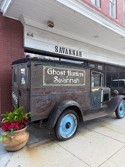 This media file shows a vintage, rusty truck parked on a sidewalk in front of a building. The truck has blue rims and a weathered, dark green body. On the side of the truck, white lettering reads "Ghost Hunters of Savannah" inside a decorative border, and below that, "GHS". A QR code is also visible on the truck's side. Next to the truck is a large terracotta pot filled with red flowers and green foliage.

The building behind the truck has white siding with vertical lines and red brick at the base. A sign on the white siding reads "SAVANNAH" in large black letters. The building number "416" is also visible. The scene appears to be outdoors on a sidewalk made of concrete tiles. The lighting suggests it could be daytime, possibly overcast given the lack of harsh shadows. The overall impression is one of a quirky, possibly historical, commercial establishment in Savannah, Georgia. No people are visible in the image.