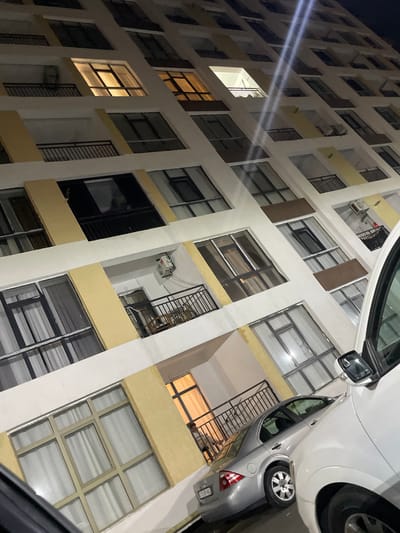 The image shows a nighttime view of a residential building in Sumgayit, Azerbaijan. The building is tall and appears to be made of concrete with yellow and white accents. Many windows are visible, some illuminated from within, suggesting occupancy. Balconies with black railings are present on several floors. In the foreground, the rear of a silver sedan is parked next to a white vehicle, which is partially visible on the right side of the frame. The lighting suggests it is after dark, with artificial lights from inside the building and potentially streetlights illuminating the scene. The overall impression is of an urban residential environment.