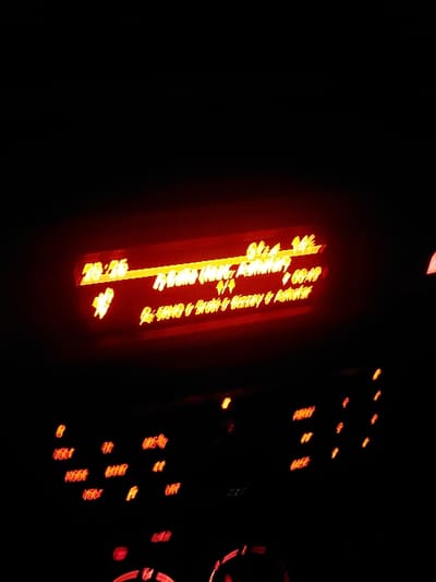 The image displays a dark interior scene, likely within a vehicle, dominated by the red and orange glow of a digital display and control panel, suggesting it is nighttime. The central element is a rectangular, red-orange digital screen, tilted slightly to the right, which shows prominent text. In the bottom-left corner of this display, the time "20:25" (8:25 PM) is clearly visible. The main text across the screen reads "TRADITIONNEL MAROCAINE," indicating "Traditional Moroccan," followed by "Majmaa Toba, Maroc," explicitly identifying Majmaa Tolba, Morocco, as a location reference or origin. A smaller "00:00" appears in the top-right of the screen, possibly a duration counter.

To the right of the main display, several smaller, scattered red and orange lights are visible, forming what appears to be a control panel. Some of these illuminated buttons feature partially legible text, such as "MODE," "BAND," "MUTE," and "SCAN," typical of an in-car audio system or media player. The overall scene suggests the vehicle's infotainment system is active, potentially playing Moroccan traditional music related to Majmaa Tolba. No people are visible in the frame, and the entire setting emphasizes artificial light against deep darkness. The image exhibits a slight blur, particularly on the smaller illuminated elements.